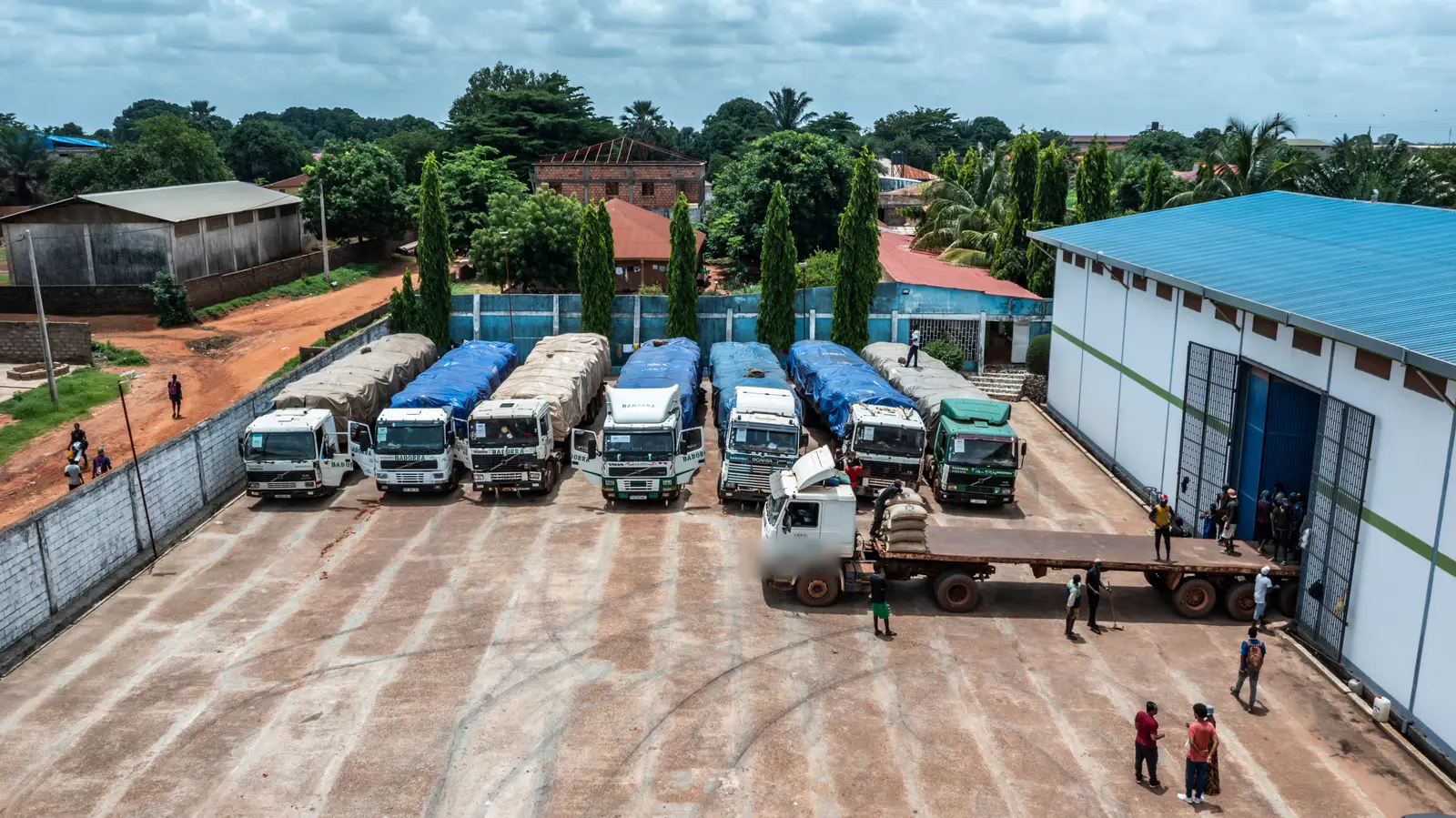 An aerial view of the depot yard with a row of sheeted trailers parked in line and a flatbed being loaded at the warehouse door.