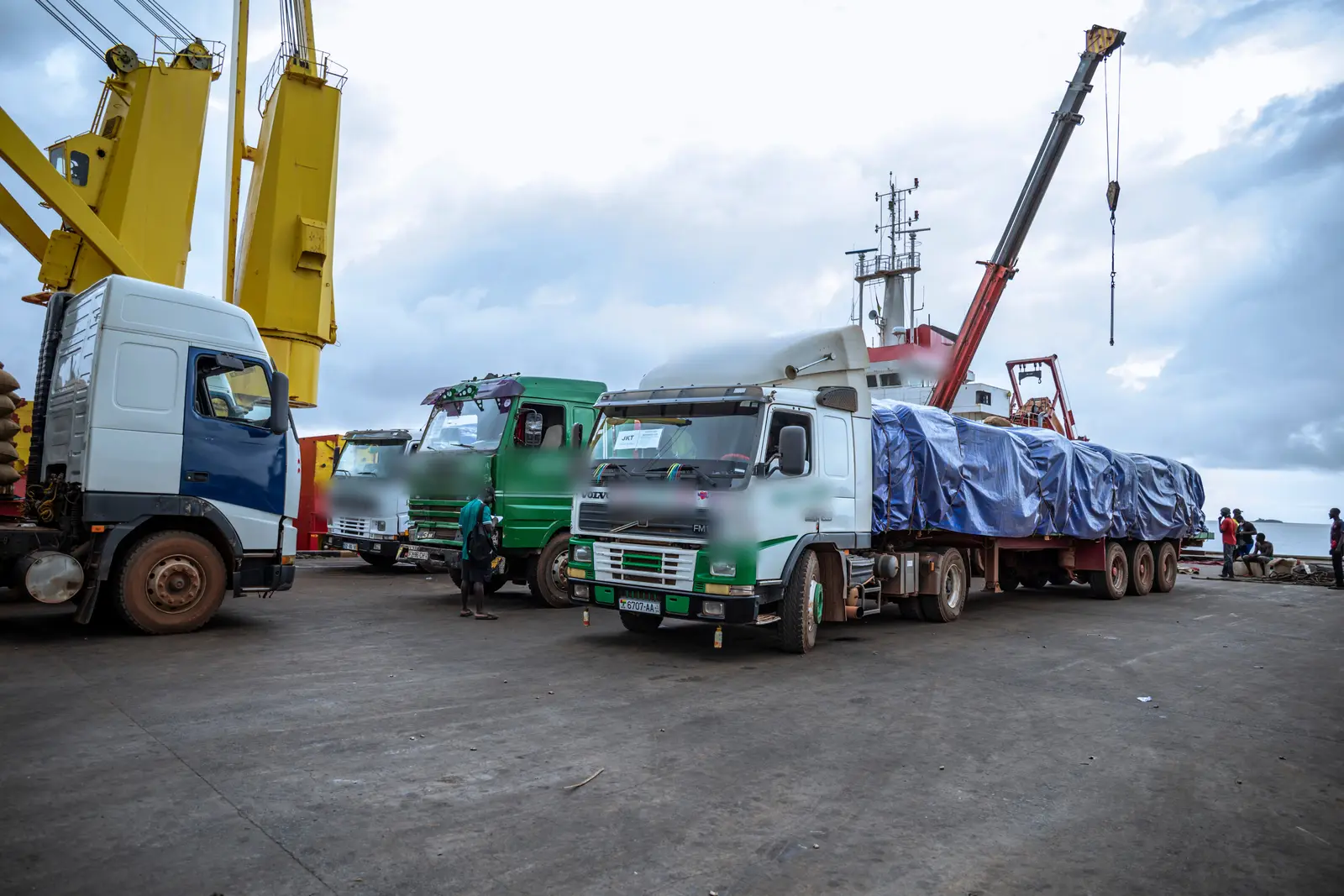 A line of loaded trucks waiting on the quay at Bissau beside the harbour cranes and the vessel.